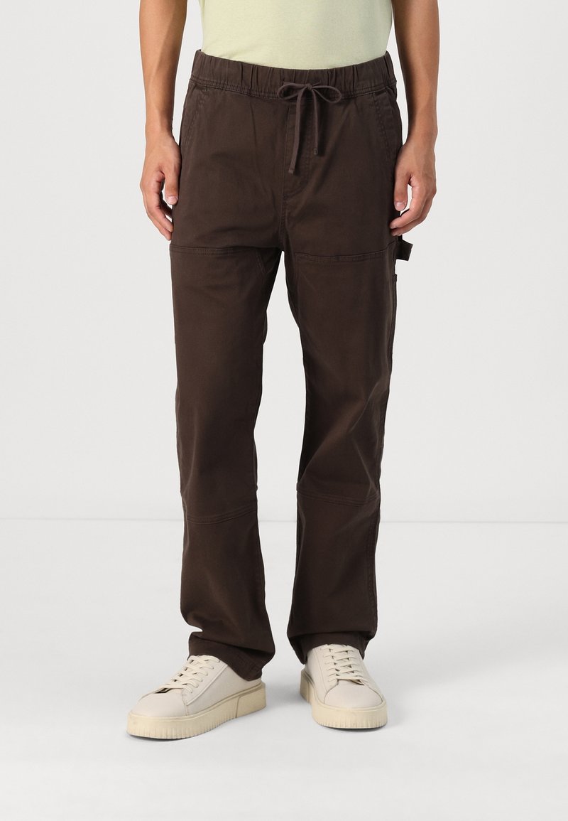 Bruna cargobyxor med elastisk midja och dragsko, med sidofickor och avslappnad passform, i kombination med ljusa sneakers.