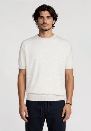 Giovane uomo con capelli scuri di media lunghezza che indossa una maglietta a maniche corte bianca tinta unita in maglia e pantaloni scuri con coulisse su sfondo semplice.