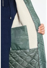 Grön vattentät jacka med quiltat mönster, pälsfoder och en ficka på sidan som visar skötselråd i vit text.