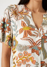 Kurzärmlige Bluse aus leichtem Stoff mit einem floralen Muster in Orange-, Blau- und Grüntönen auf weißem Hintergrund.