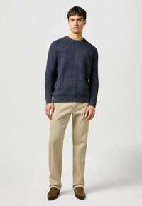 Marinblå stickad tröja med ribbad nederkant och ärmar, tillsammans med ljus khaki raka byxor och bruna skor, stående mot en neutral bakgrund.