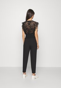 Black lace top with ruffled sleeves and a fitted waist, paired with black tapered pants and white sneakers against a plain background.