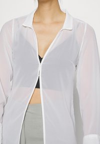 Camisa blanca transparente con diseño de cuello y escote en V bajo, con mangas largas y cierre de cremallera blanca contrastante.