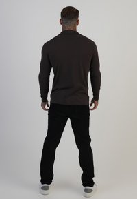 Cămașă cu mânecă lungă, de culoare maro închis, dintr-un material neted, purtată cu pantaloni negri și pantofi albi. Tatuajele vizibile pe spate adaugă detalii.