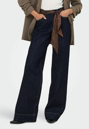 Femme portant un jean bleu foncé à jambes larges avec une ceinture en foulard marron à motif, les mains dans les poches et des chaussures noires pointues.
