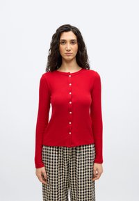 Γυναίκα με σγουρά σκουρόχρωμα μαλλιά φοράει κόκκινο μπλουζάκι με κουμπιά και μακριά μανίκια και ασπρόμαυρο καρό παντελόνι, στέκεται μπροστά σε απλό λευκό φόντο.