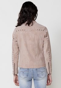 Jachetă din imitație de piele bej deschis, cu accente cu bolțuri pe umeri și mâneci. Prezintă un design ajustat și manșete cu fermoar.