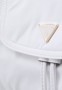 Mochila de nylon blanca con un logo de hardware dorado en forma de triángulo. Presenta un cierre de solapa y cuerdas texturizadas para ajuste.