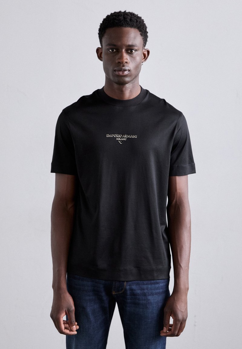 T-shirt noir à manches courtes et col rond en tissu lisse, avec un petit logo brodé à l'avant, associé à un jean bleu.