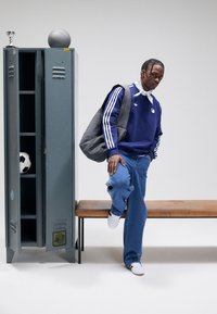 Μπλε φούτερ με λευκές ρίγες, γκρι τσάντα και μπλε τζιν. Ξύλινος πάγκος κοντά σε γκρι ντουλάπι. Μπάλα ποδοσφαίρου και τρόπαιο μέσα στο ντουλάπι.