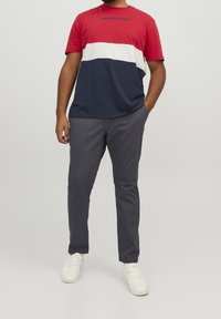 Tričko s pruhy v červené, bílé a námořnické modři, se znakem "JACK & JONES"; nošeno s šedými kalhotami a bílými teniskami. Uvolněný střih a krátké rukávy.