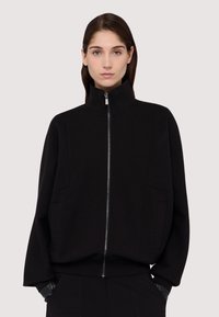 Černá zipová bunda vyrobená z měkkého materiálu, s vysokým límcem, bočními kapsami a širokými rukávy pro uvolněný střih.