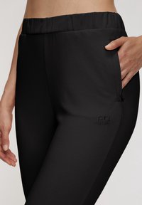 Black athletic pants featuring a stretchy waistband, side pocket, and embroidered logo detail on the thigh. Smooth fabric texture.