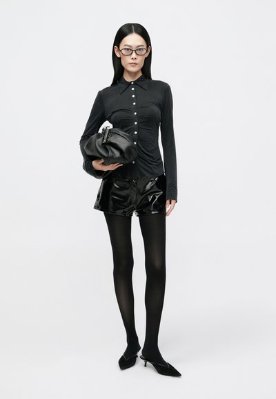 Black long-sleeve top with button details, paired with shiny black patent shorts and opaque black tights. Carrying a black textured bag.