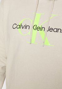 Felpa beige in cotone con un logo nero e verde neon di grandi dimensioni. Include coulisse bianche con estremità in metallo per la regolazione.