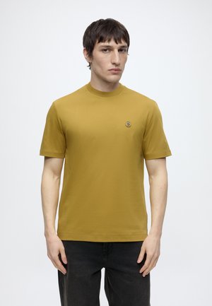 Mand med kort mørkt hår iført en sennepsgul kortærmet skjorte med lille rundt logo på brystet og sorte bukser, står foran en ensfarvet baggrund.