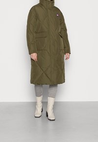 Cazadora acolchada verde oliva hasta la rodilla con cuello alto, patrón de rombos, dos bolsillos delanteros y logo de la marca en el pecho.