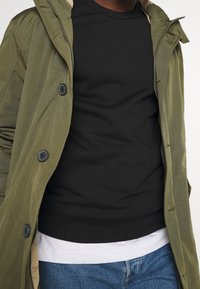 Parka verde oliva con bottoni e cerniera, indossata sopra una felpa nera. Orlo della camicia bianca visibile e jeans blu. Texture del tessuto liscia.