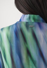 Blusa di seta con un motivo astratto verde e blu, dotata di colletto e di un piccolo logo metallico sulla parte posteriore del collo. Tessuto morbido e liscio.