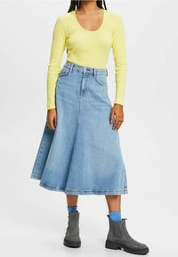 Gelbes, geripptes Langarmoberteil kombiniert mit einem blauen Jeans-A-Linienrock, der eine hohe Taille und einen ausgestellten Saum hat. Graue Stiefeletten sind unten sichtbar.