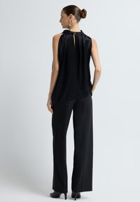 Zwarte mouwloze top met een keyhole-detail aan de achterkant, gecombineerd met zwarte op maat gemaakte broeken. Glad materiaal en een ontspannen pasvorm met een lichte glans.