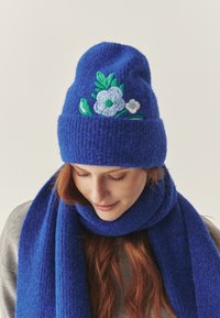 Căciulă tricotată albastră cu un finisaj texturat, având broderie florală în nuanțe de albastru și verde. Asortată cu o eșarfă albastră corespunzătoare.