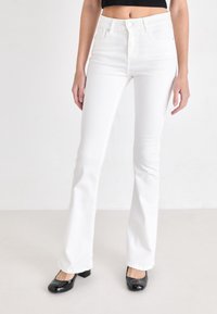 Jeans flared blancos con cintura alta, hechos de mezclilla. Cuenta con un diseño de cinco bolsillos y cierre de botón. Combinados con zapatos negros de tacón cuadrado.