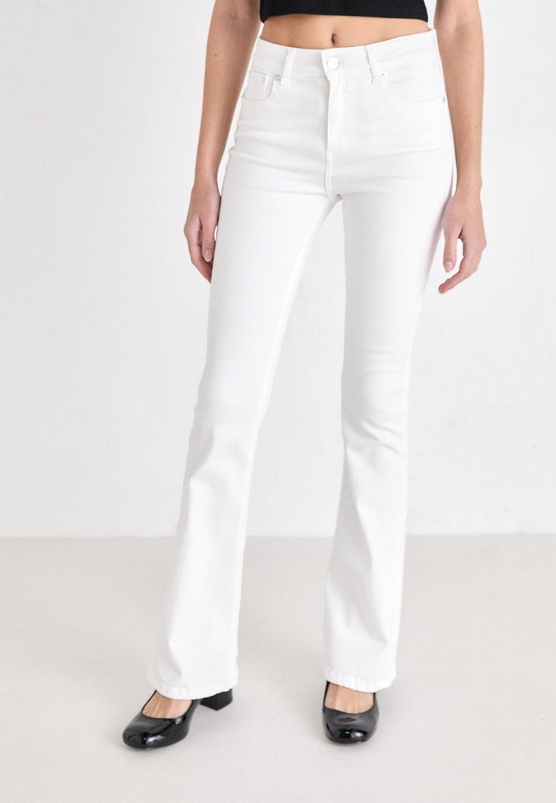 Jeans flared blancos con cintura alta, hechos de mezclilla. Cuenta con un diseño de cinco bolsillos y cierre de botón. Combinados con zapatos negros de tacón cuadrado.