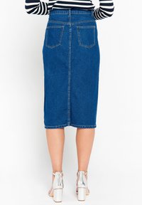 Falda lápiz de mezclilla en azul medio con corte recto, bolsillos traseros y una costura visible que corre por el centro, combinada con tacones plateados.
