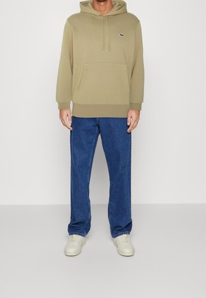 Beige huvtröja med känguruficka och dragsko. Kombinerad med blå jeans och vita sneakers. Diskret logotyp på bröstet. Mjuk textur.