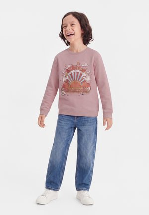 Enfant aux cheveux foncés souriant et debout, portant un sweat-shirt rose Stella McCartney, un jean bleu et des baskets blanches.