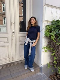 Tengerészkék póló "love" felirattal, világosszürke pulóver derékban megkötve, magasderekú farmernadrág, világos színű cipők. Szabadtéri környezet.