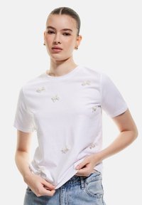 Camiseta blanca de algodón con mangas cortas y cuello redondo, que presenta detalles bordados de lazo en hilo blanco en la parte frontal.