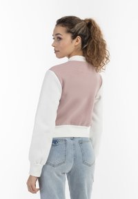 Helles rosa Bomberjacke mit weißen langen Ärmeln und geripptem Saum. Weiche Textur mit Rundkragen, getragen über hellblauen Jeans.