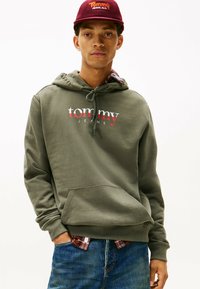 Felpa verde oliva con tasca frontale e cappuccio con coulisse, con la scritta "tommy" e "JEANS" in testo bianco e rosso. Indossata sopra una camicia a quadri.