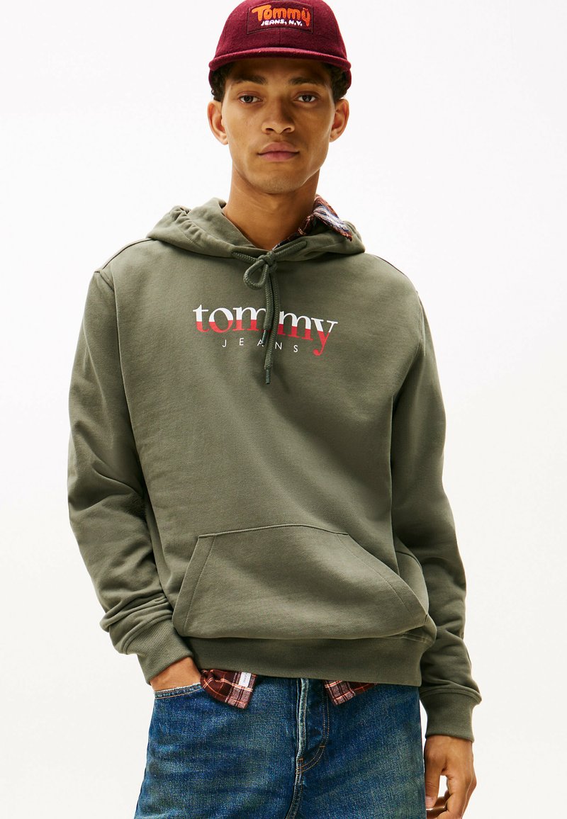 Felpa verde oliva con tasca frontale e cappuccio con coulisse, con la scritta "tommy" e "JEANS" in testo bianco e rosso. Indossata sopra una camicia a quadri.