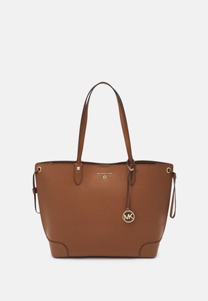 Brązowa skórzana torba typu tote z podwójnymi uchwytami, złotą zawieszką z logo „MK” i oznaczeniem Michael Kors na jednolitym jasnym tle.
