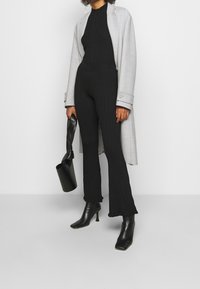 Jumpsuit en maille côtelée noire avec des jambes évasées, associé à un long manteau gris clair. Porte un sac à main noir. Bottes à talons noires.