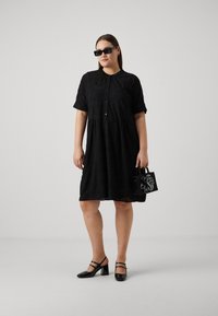 Μαύρο φόρεμα κοντό μανίκι με κουμπιά μπροστά και υφή. Συνδυασμένο με μαύρες λουστρίνι ψηλές γόβες και μια μικρή μαύρη τσάντα με λεπτομέρεια φιόγκου.