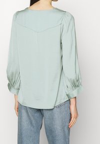 Hellgrüne Satinbluse mit einem Rückennähten, puffweiten Ärmeln und Manschetten. Glatte Textur, lockere Passform, kombiniert mit hellblauen Jeans.