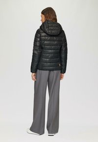 Doudoune noire à capuche, avec des panneaux matelassés, portée avec un pantalon gris à jambes larges. Design simple avec des accents minimalistes.