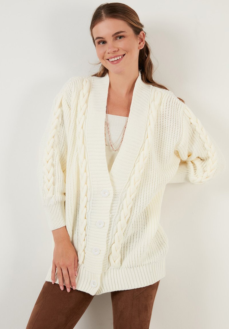 Cárdigan blanco oversized con escote en V, textura trenzada en las mangas y botones grandes. Se lleva sobre una blusa más clara con un acabado suave.