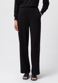 Pantalones amplios negros de tejido suave, con una cintura alta y bolsillos laterales, combinados con sandalias negras de tacón abierto.