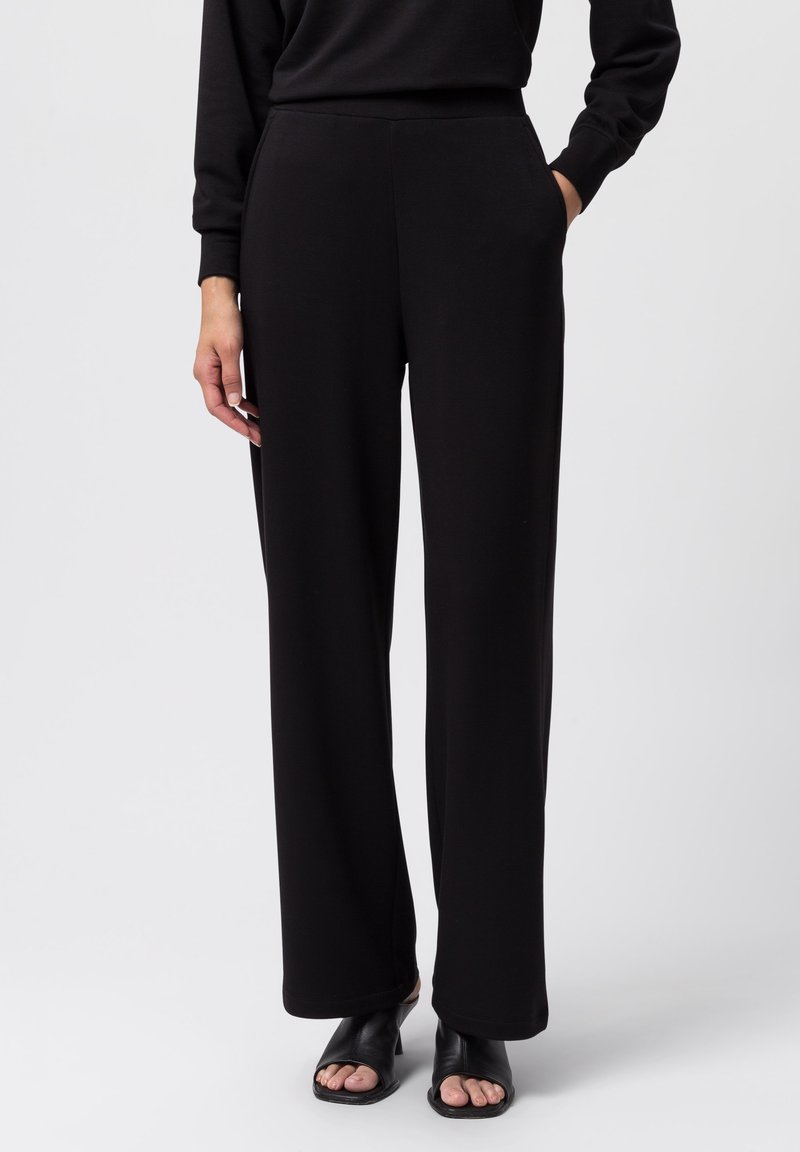 Pantalones amplios negros de tejido suave, con una cintura alta y bolsillos laterales, combinados con sandalias negras de tacón abierto.