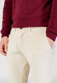 Pantalones de pana beige con textura acanalada vertical, corte slim, con bolsillos delanteros y cierre con botón negro. Combinados con un suéter burdeos.