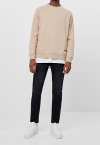 Beige sweatshirt med rund halsringning, ribbade ärmslut och nederkant, lager-på-lager över en vit skjorta. Bärs med mörka jeans och vita sneakers.