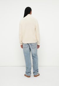Maglione lavorato a maglia di colore crema con colletto alto, abbinato a jeans larghi di colore blu chiaro e sneakers a fantasia. Texture morbida e vestibilità rilassata.