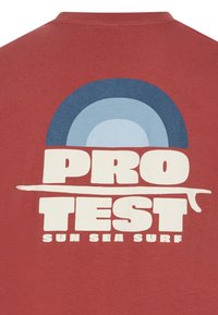 Bordo marškinėliai su dideliu grafiniu dizainu, kuriame yra koncentriški mėlyni apskritimai ir ryškus tekstas "PRO TEST" bei "SUN SEA SURF" baltos spalvos.