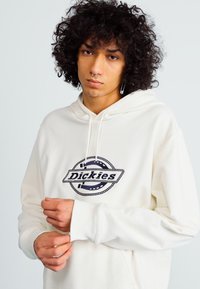 Kremfarget hettegenser med stor svart brodert "Dickies"-logo og kontrasterende designdetaljer. Myk stoff med en avslappet passform.