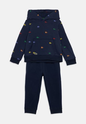 Tumši zila kapucīna krekla un bikses komplekts ar daudzkrāsainām izšūtām krokodila emblēmām, kas izkaisītas pa krekla virsmu.